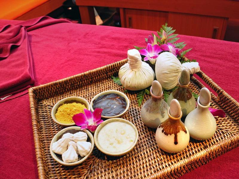 Tablett mit verschiedenen Kräutern, Pulver, Cremes und dekorativen Blüten auf rotem Tuch.