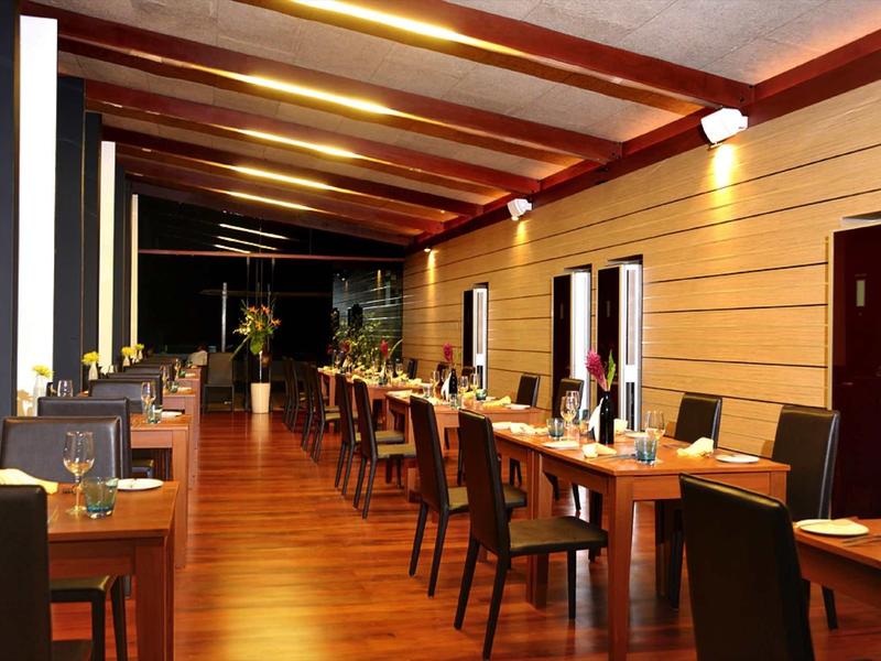 Moderner Speisesaal mit Holzboden, roten Stühlen und langen Tischen unter warmem Deckenlicht.