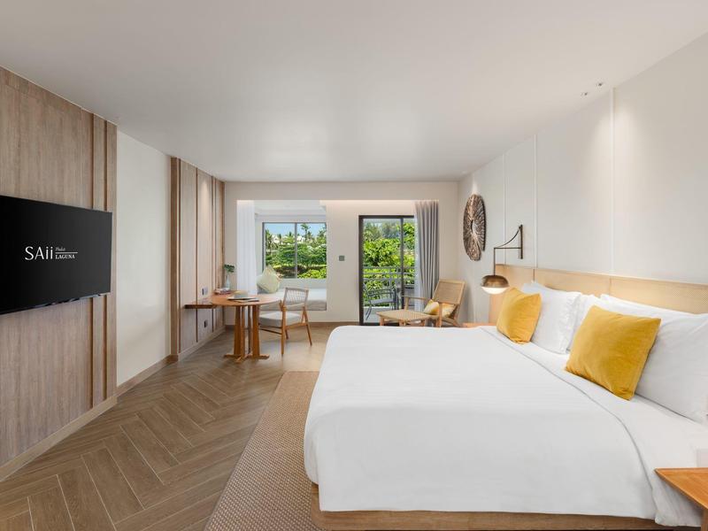 Modern hotelkamer met tweepersoonsbed, gele kussens, bureau en uitzicht op een groen terras.