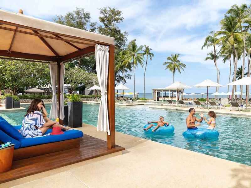 Ontspannen mensen in het zwembad en op een ligstoel onder een overdekte cabana met tropische omgeving.