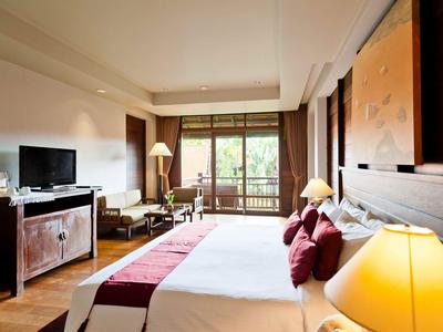 Helles Hotelzimmer mit großem Bett, roten Kissen, Fernseher, Sessel und Balkon mit Holzgeländer.