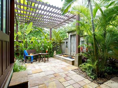 Gemütlicher Garten mit Holzpergola, Steinboden, Pflanzen und Sitzbereich mit blauem Stuhl.