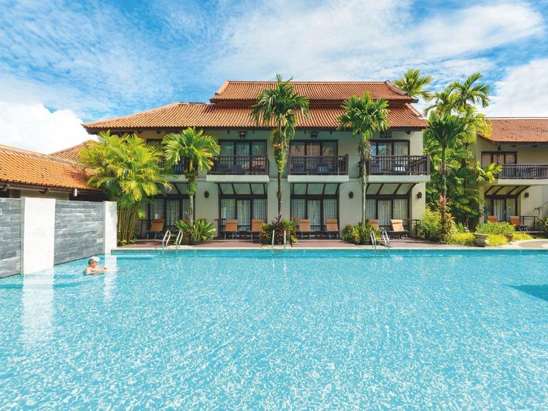 Hotel met groot zwembad, tropische planten en heldere blauwe lucht.