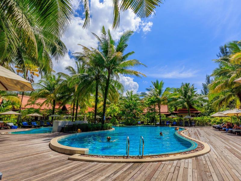 Rond zwembad met waterfonteinen, omgeven door houten dek, palmbomen en parasols onder een blauwe lucht.