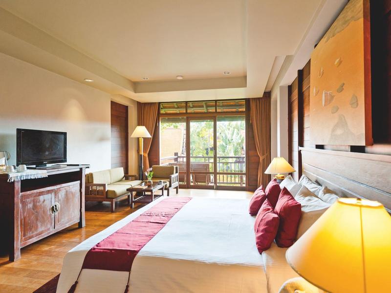 Lichte hotelkamer met groot bed, tv, zithoek en balkon met uitzicht op groen.