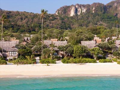 Beach with white sand, palm-surrounded bungalows, and forested mountains in the background