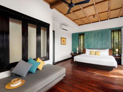 Bright hotel room with wooden floor, large windows, double bed, and sofa with colorful cushions.