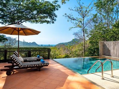 Pool area with lounge chairs, umbrella, and view of green hills and sea in the background