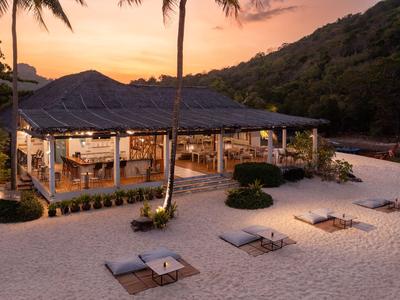 Offenes Strandrestaurant bei Sonnenuntergang mit Tischen im Sand und Palmen im Hintergrund.