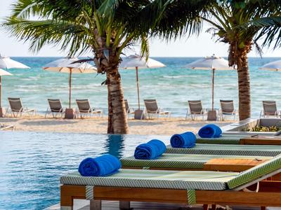 Poolbereich mit Liegestühlen, blauen Handtüchern und Blick auf Palmen und Strand.