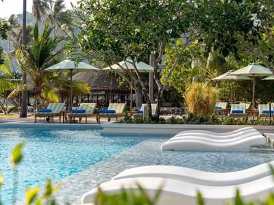 Ein Swimmingpool mit weißen Liegestühlen und Sonnenschirmen neben üppigem Grün im Urlaubsresort.