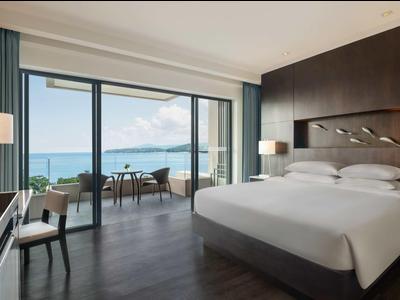 Camera d'hotel moderna con letto grande, area salotto e vista sul mare attraverso finestre dal pavimento al soffitto.