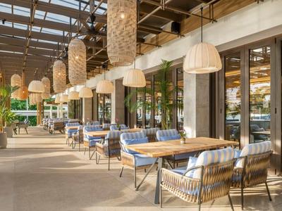 Ristorante all'aperto con tavoli in legno, sedie con cuscini e grandi lampade sospese sotto un tetto di vetro.