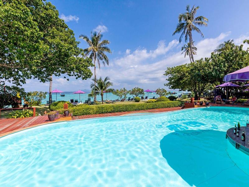 Clear water pool with palm trees and a view of the beach and sea under a sunny sky.