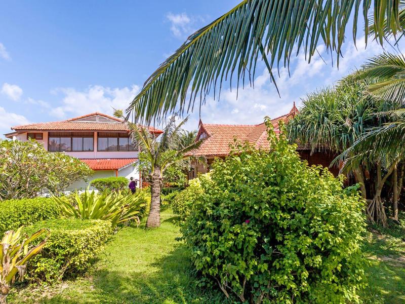 Hotel building surrounded by lush tropical garden with palm trees and green bushes under blue sky.