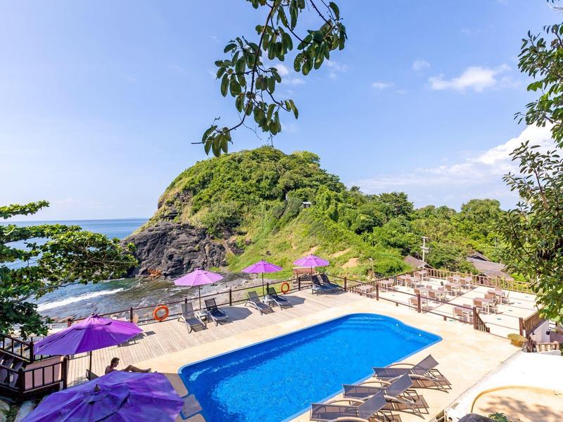 Hotel pool with lounge chairs and umbrellas near a tropical coastal hill under a blue sky.