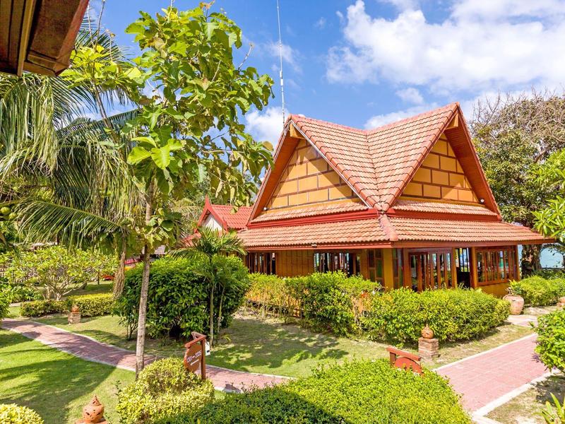 Traditional wooden house with red roof surrounded by tropical garden and blue sky.