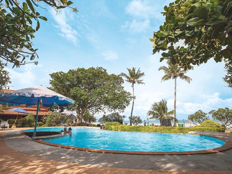 Round pool with sun umbrellas and trees under blue sky in a tropical resort.