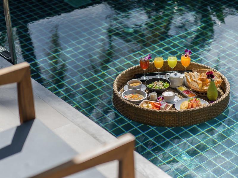 Floating breakfast tray with various foods and drinks in pool next to chairs.