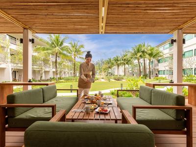 Osoba připravuje jídlo na dřevěném stole pod zastřešenou terasou s výhledem na zahradu s palmami.