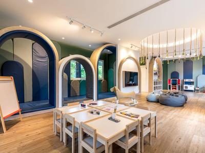 Moderner Kinderspielraum mit Holztischen, Stühlen und bunten Podesten unter großen Rundbögen.