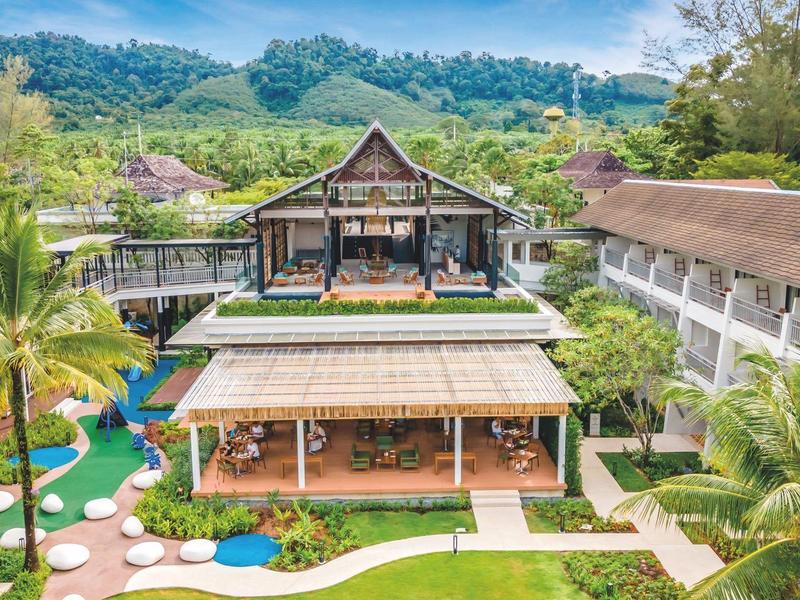 Ein tropisches Resort mit Holzgebäuden, grünen Gärten und umgeben von Hügeln unter blauem Himmel.