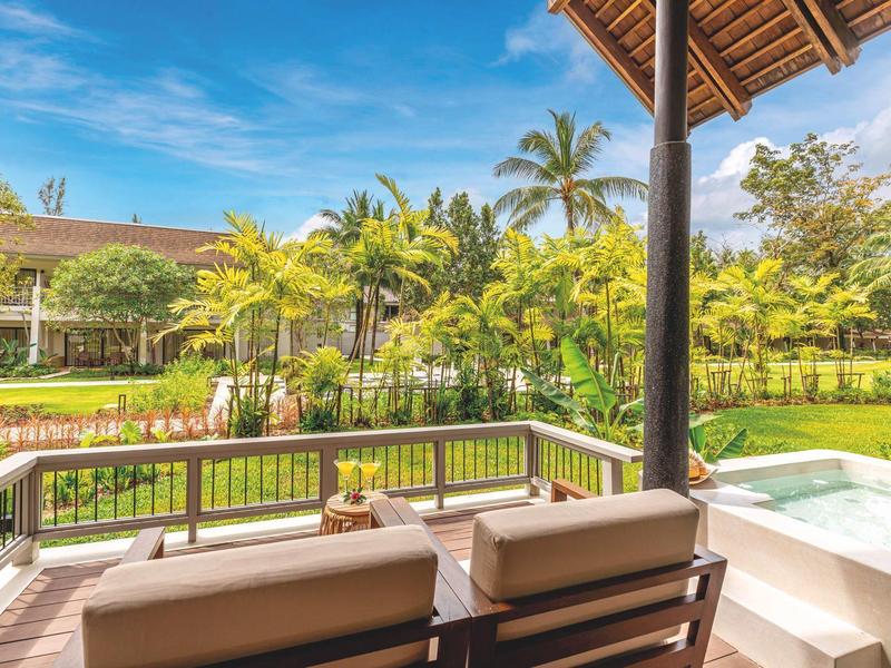 Blick von einer Terrasse mit Sitzbank und Whirlpool auf tropischen Garten bei sonnigem Himmel.