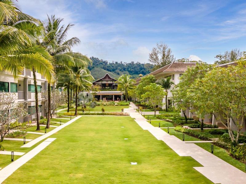 Ein gepflegter Garten mit palmengesäumtem Weg zwischen Hotelgebäuden unter blauem Himmel.