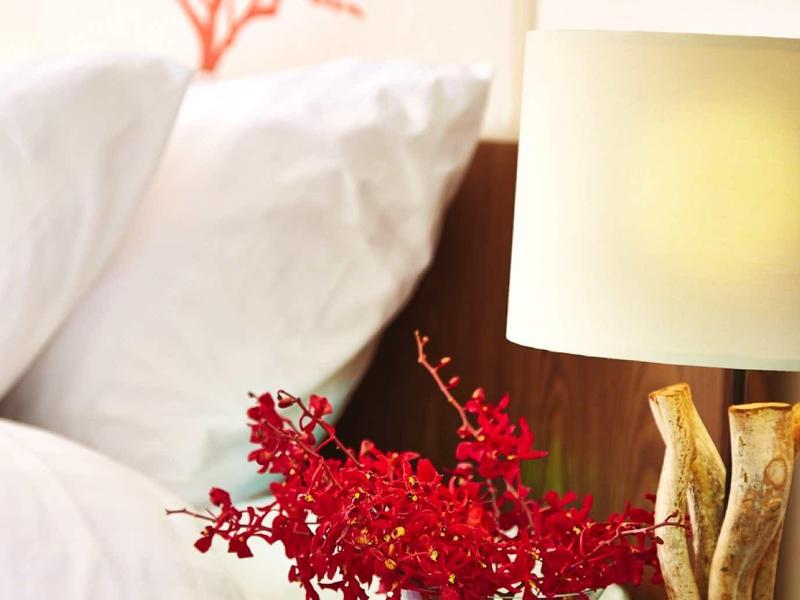 Rotes Blumenarrangement in Glasvase neben Holzlampe auf Nachttisch mit weißem Bett und Wanddeko.