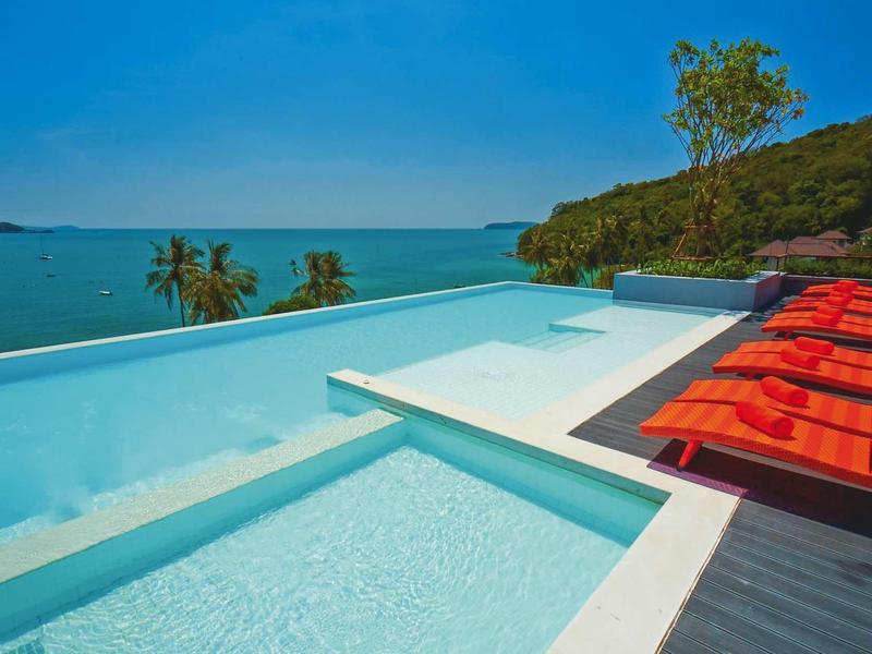 Moderner Pool mit klarem Wasser, roten Liegestühlen und Meerblick unter blauem Himmel.