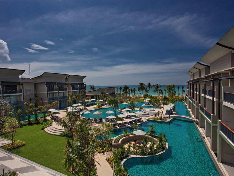 Modern hotelcomplex met zwembad, ligstoelen en uitzicht op de zee bij heldere lucht.