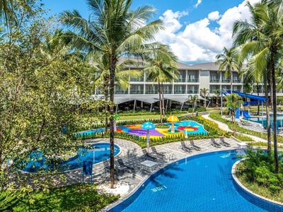 Hotelzwembad met blauwe glijbaan, ligstoelen en tropische tuin bij zonnige lucht.