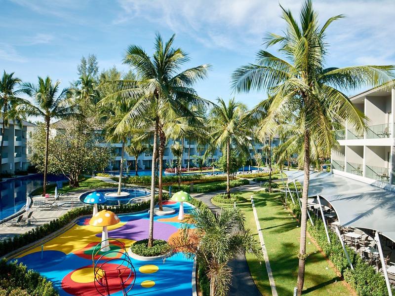 Hotel tuin met palmbomen, kleurrijke speeltuin voor kinderen en ligstoelen onder blauwe lucht.