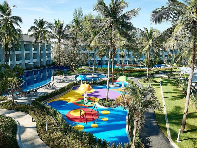 Uitzicht op een hotelzwembad met waterspeeltuin, omgeven door palmbomen en groene zones.