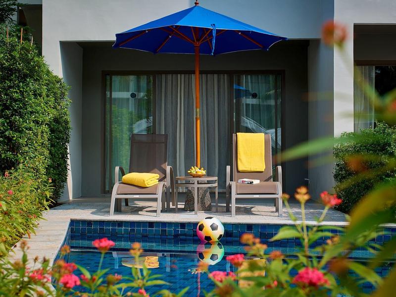 Zwembad met twee stoelen, blauwe parasol en gele kussens voor een modern gebouw.