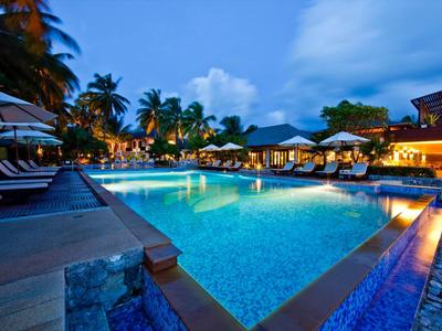 Verlichte zwembadruimte van een resort met palmbomen en ligstoelen bij schemering.