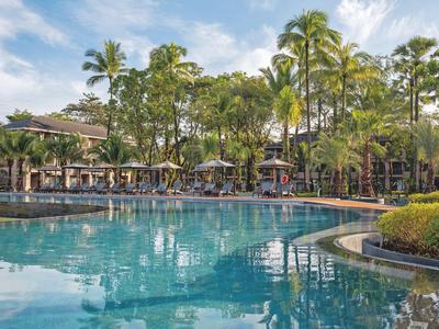 Duży hotelowy basen z leżakami i parasolami otoczony palmami i budynkami.