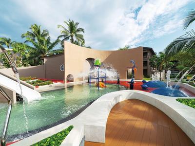 Modern buitenzwembad met waterspeeltoestellen en glijbanen, omgeven door palmbomen en een gebouw.