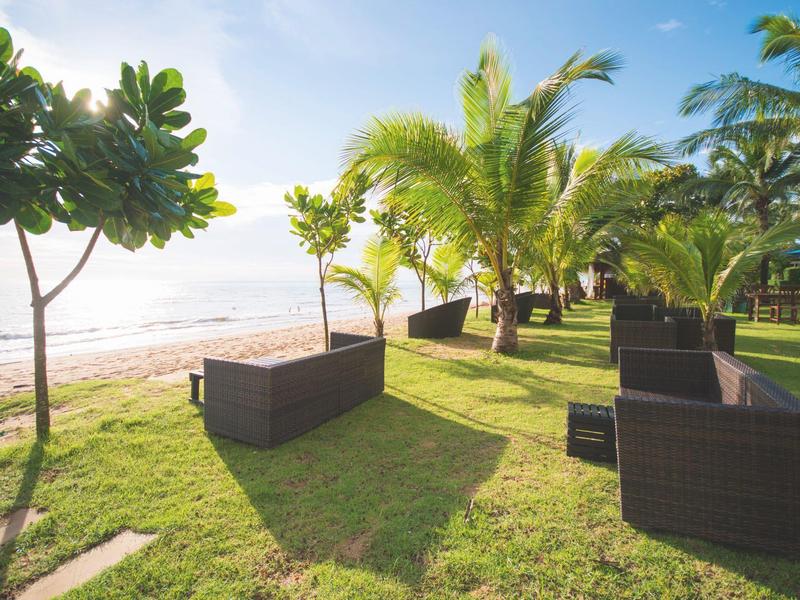 Buitenbanken op een groen gazon met uitzicht op een zonnig strand met palmbomen.