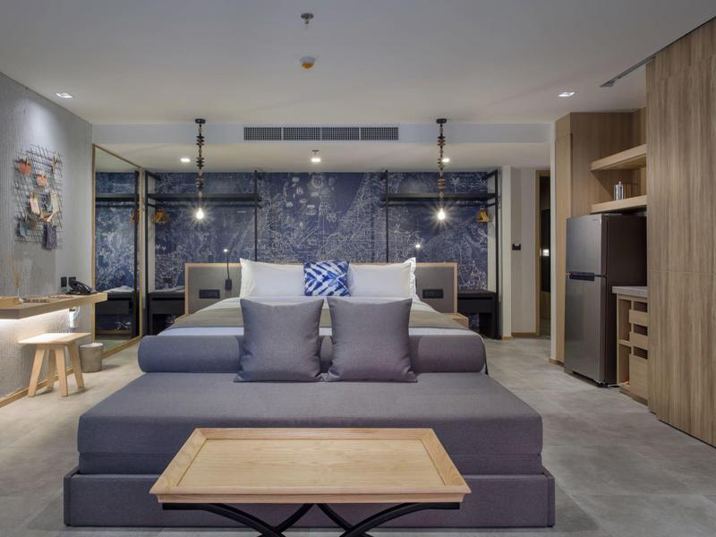 Modern furnished hotel room with double bed, sofa, and wooden furniture in neutral tones.