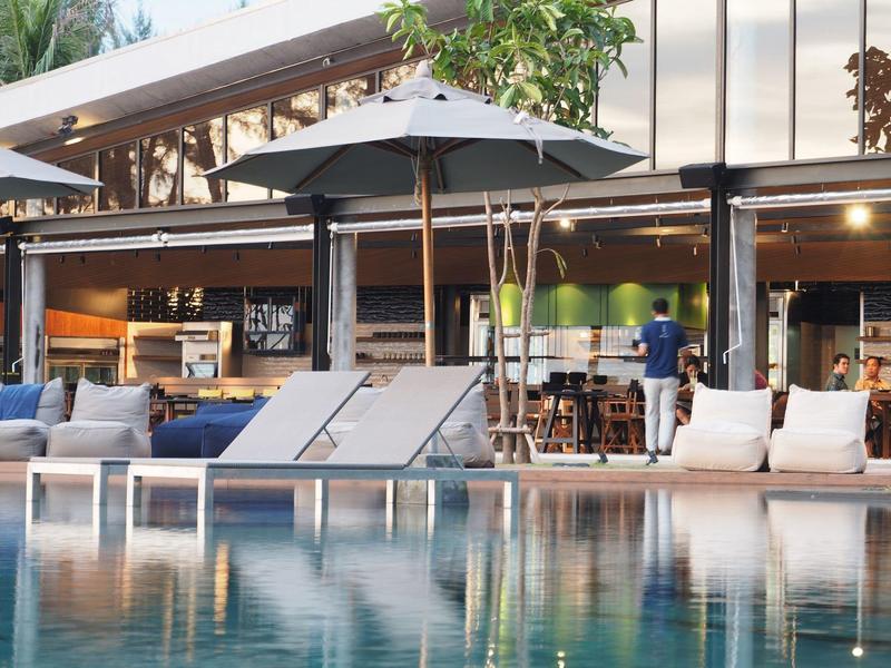 Modern hotel pool lounge with sunbeds and umbrellas in front of an open-designed building.
