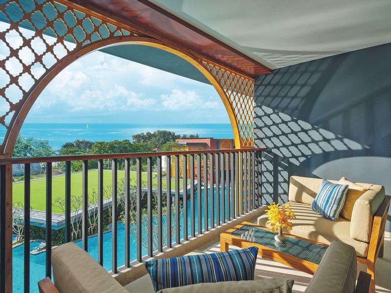 Balcón con muebles y vistas al mar, piscina y césped en un hotel tropical.