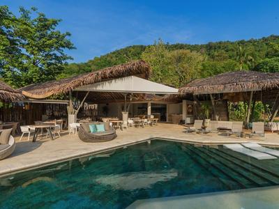 Area piscina con lettini e tetti di paglia davanti a un edificio dell'hotel, circondato da colline verdi.