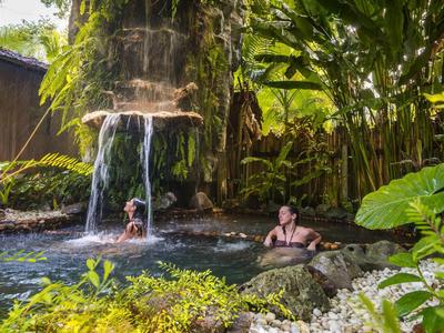 Dos personas descansan en una piscina natural con cascada rodeada de vegetación exuberante.
