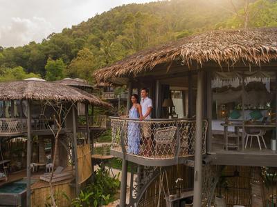 Una pareja está en el balcón de una cabaña de madera en la montaña rodeada de naturaleza verde.