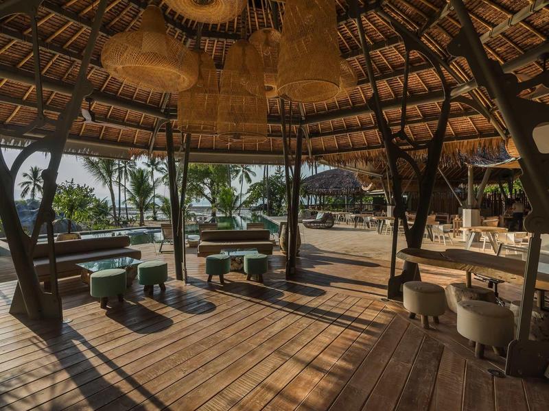 Padiglione aperto sulla spiaggia con pavimento in legno, sedute e vista su palme e mare.