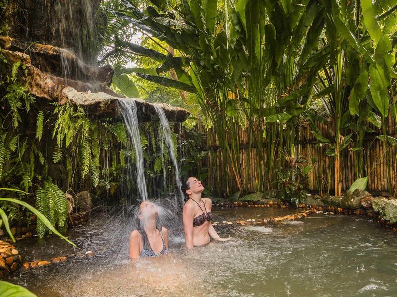Due persone si rilassano in una piscina naturale sotto una piccola cascata circondata da vegetazione rigogliosa.