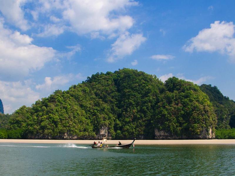 Barca a coda lunga che naviga su acqua calma davanti a un'isola boscosa sotto un cielo azzurro.