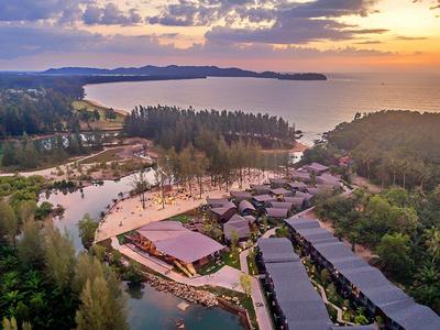 Letecký pohled na resort s bazénem, obklopený lesem a výhledem na moře za západu slunce.