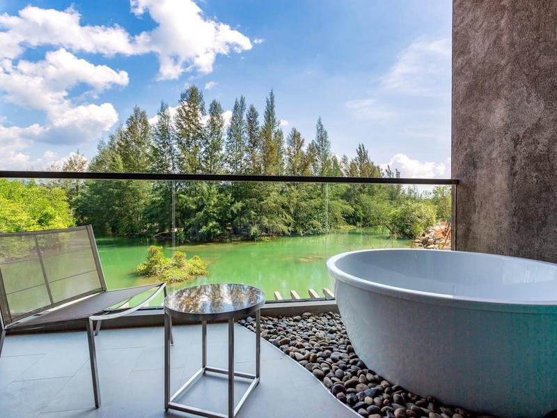 Volně stojící vana a židle na balkoně s výhledem na zelenou louku a stromy pod modrou oblohou.